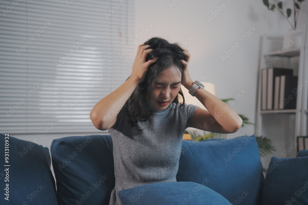 Young woman is sitting on her sofa, holding her head in her hands, suffering from a headache Young woman is sitting on her sofa, holding her head in her hands, suffering from a headache