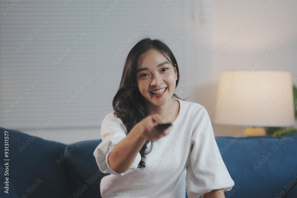 Young woman is sitting on a blue sofa in her living room, smiling and using a remote control Young woman is sitting on a blue sofa in her living room, smiling and using a remote control