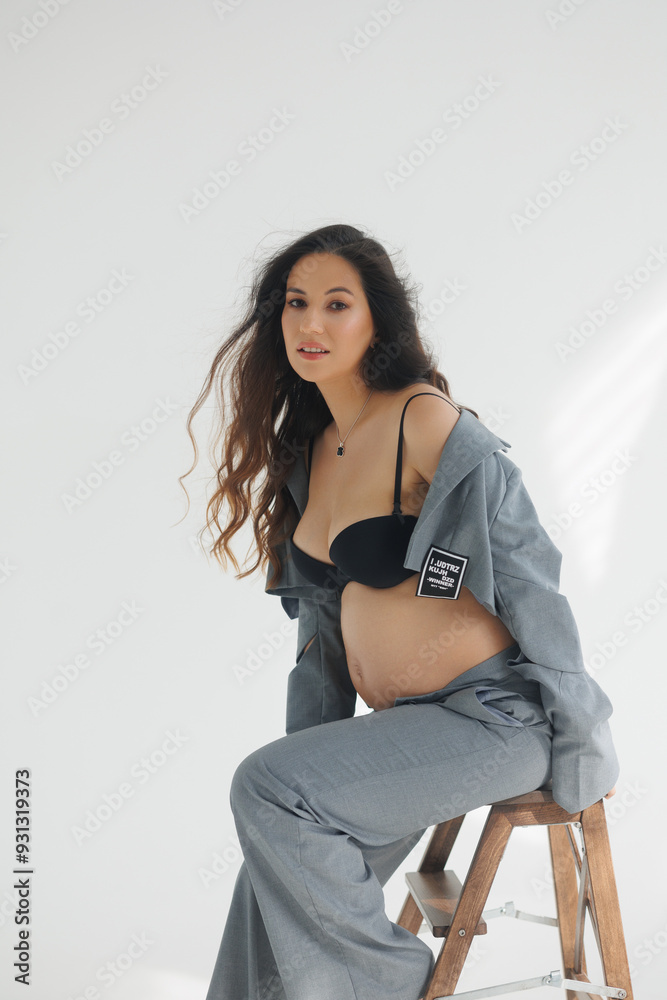 A beautiful pregnant brunette woman in a cropped gray stylish suit on a white background in the studio. A pretty girl with a bare stomach in a fashionable suit is sitting on wooden staircase barefoot. A beautiful pregnant brunette woman in a cropped gray stylish suit on a white background in the studio. A pretty girl with a bare stomach in a fashionable suit is sitting on wooden staircase barefoot.