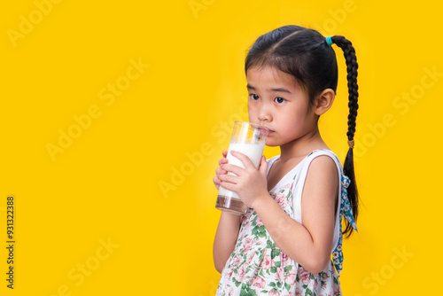 Reluctant Asian girl with pigtails holding a glass of milk, yellow background. photo