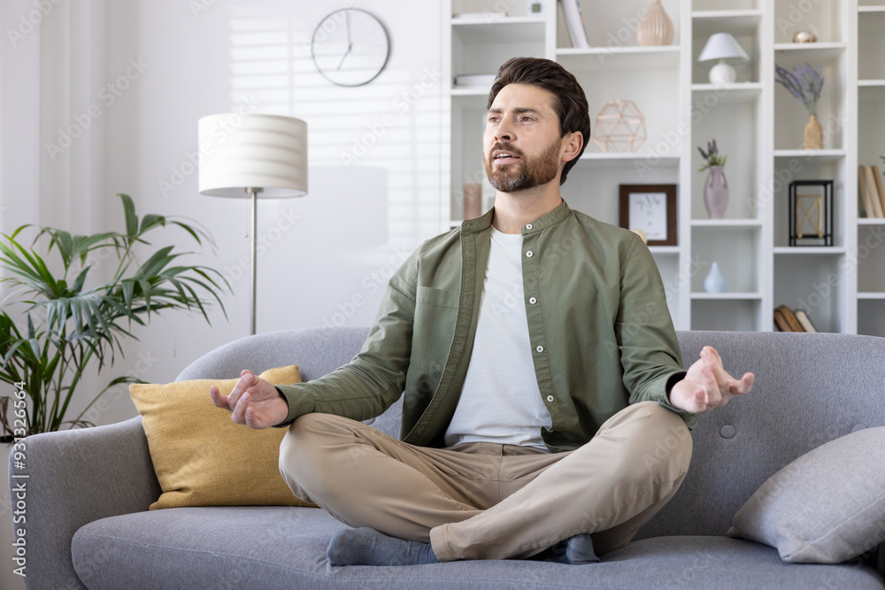 Man practicing meditation on sofa in modern living room. Embracing mindfulness and relaxation in peaceful home setting. Person sitting cross-legged, eyes closed, focused on inner peace and calm. Man practicing meditation on sofa in modern living room. Embracing mindfulness and relaxation in peaceful home setting. Person sitting cross-legged, eyes closed, focused on inner peace and calm.