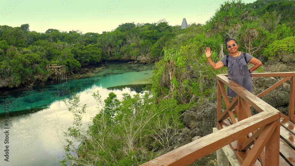 Fototapeta premium lächelnde Asiatin winkt fröhlich vom Aussichtssteg der blauen Lagune im Westen der Insel Sumba in tropischer Landschaft