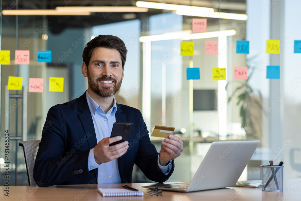 Businessman holding credit card and smartphone in office, exploring online payment and e-commerce. Man in suit demonstrating modern business environment with focus on digital finance solutions