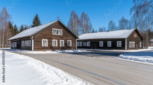 A quaint wooden church and traditional buildings are blanketed in snow, surrounded by bare trees against a vibrant blue winter sky in an ancient Russian village.