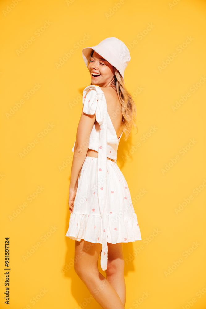 Young beautiful smiling blond female in trendy summer white top and skirt clothes. Carefree woman posing near yellow wall in studio. Positive model in panama hat. Cheerful and happy Young beautiful smiling blond female in trendy summer white top and skirt clothes. Carefree woman posing near yellow wall in studio. Positive model in panama hat. Cheerful and happy