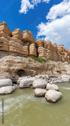 The river reflects the towering rocky mountains, while brown sand is visible along the water's edge on a sunny day, creating a picturesque natural landscape