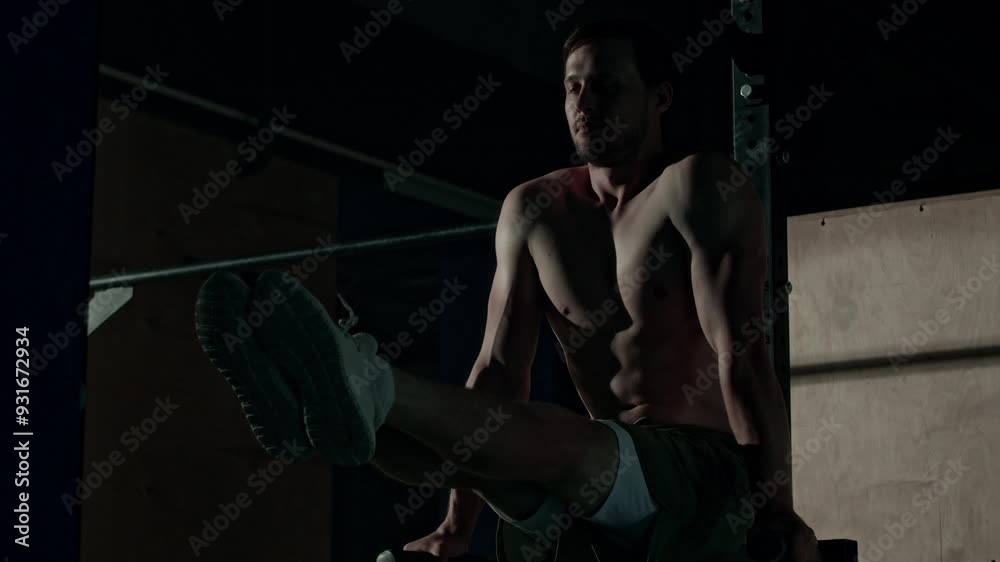 Athletic young Caucasian shirtless man doing twisting leg raise on parallel bar while training in dark at gym Athletic young Caucasian shirtless man doing twisting leg raise on parallel bar while training in dark at gym