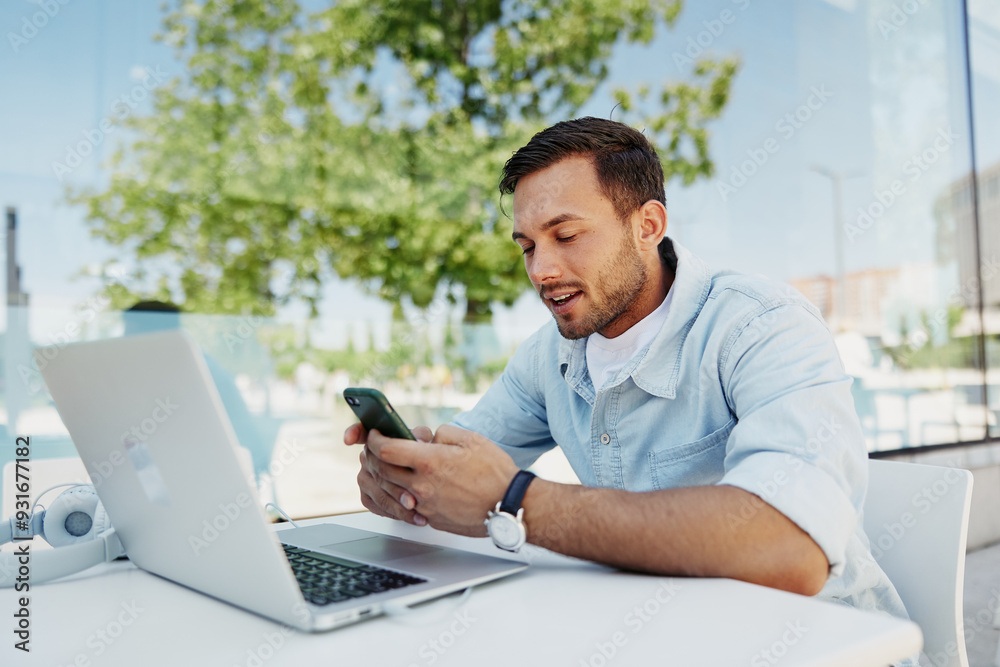 Young man smiling while checking his smartphone at an outdoor café, working on a laptop, enjoying a sunny day Young man smiling while checking his smartphone at an outdoor café, working on a laptop, enjoying a sunny day