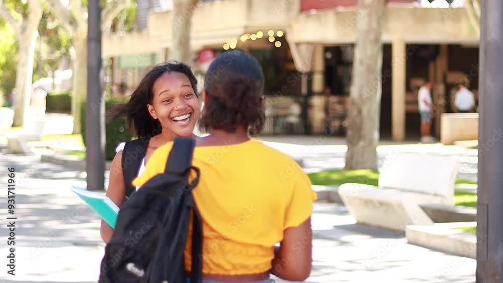 Two young Hispanic woman students meeting on the street, greeting each other and starting a conversation, carrying notebook and backpacks, one wearing headphones and holding a smartphone Two young Hispanic woman students meeting on the street, greeting each other and starting a conversation, carrying notebook and backpacks, one wearing headphones and holding a smartphone