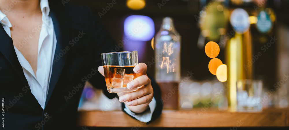 A stylish man holds a glass of whiskey in a fancy pub amidst a crowd of celebrants. The warm and elegant atmosphere accentuates the rich amber-colored liquid. A stylish man holds a glass of whiskey in a fancy pub amidst a crowd of celebrants. The warm and elegant atmosphere accentuates the rich amber-colored liquid.