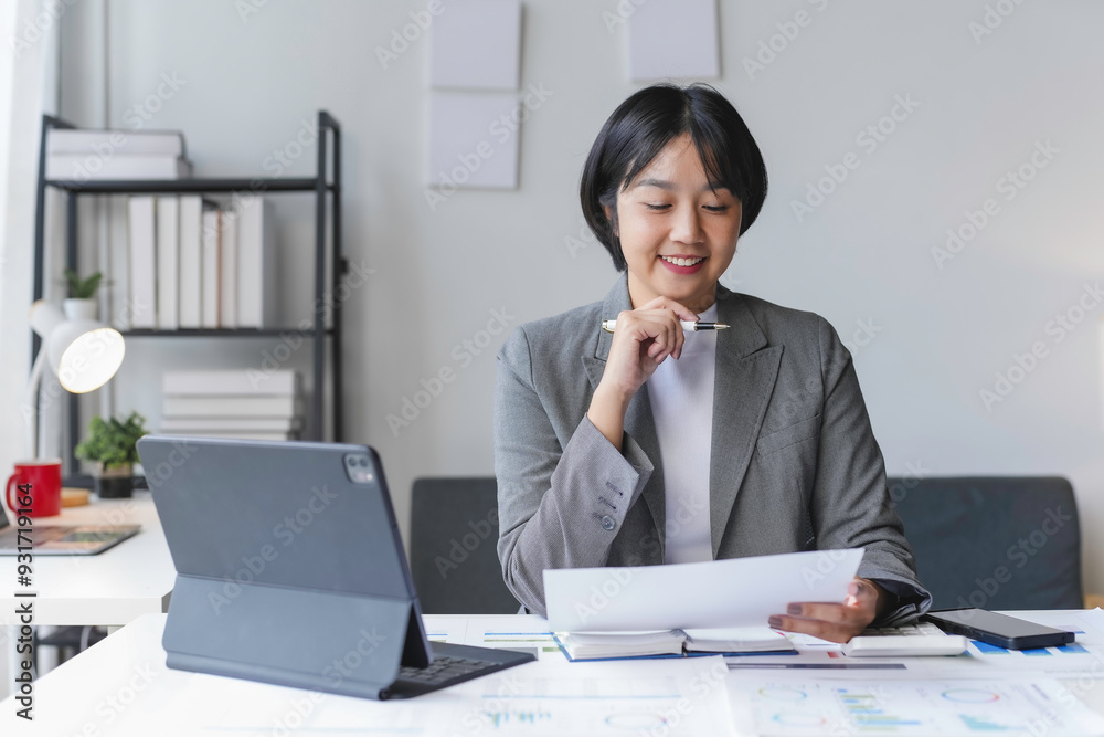 Asian businesswoman in a gray business suit smiles as she reviews documents at her desk Asian businesswoman in a gray business suit smiles as she reviews documents at her desk