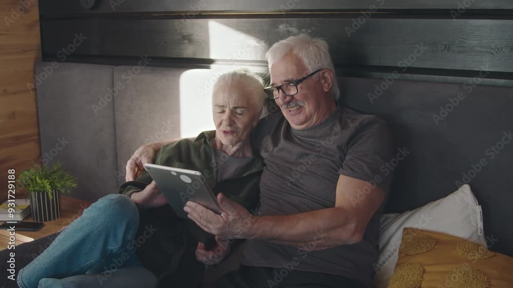 Medium shot of smiling elderly Caucasian husband and wife relaxing on bed together, hugging, looking at tablet computer screen, browsing internet, shopping online or booking vacation Medium shot of smiling elderly Caucasian husband and wife relaxing on bed together, hugging, looking at tablet computer screen, browsing internet, shopping online or booking vacation