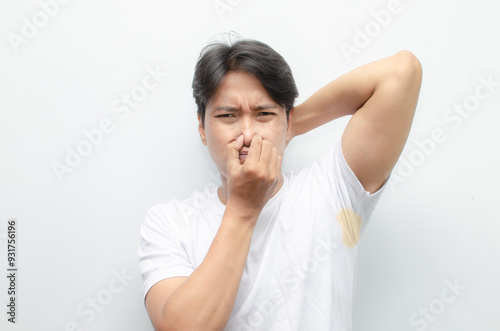 asian man feels bad after smelling his armpit with yellow stain on his white tshirt. young asian man smells his bad odor armpit with disgusted face.