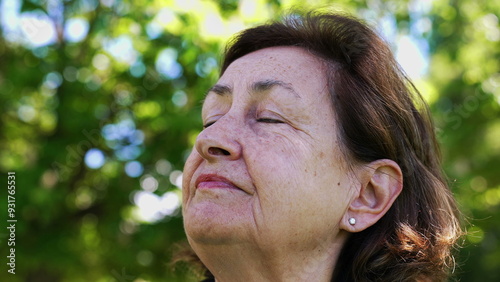 Contemplative Elderly woman with eyes closed, enjoying the warmth of the sun in a park. green leaves create a tranquil and peaceful atmosphere, opening eyes in meditation photo