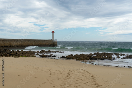 Au bout du môle se dresse le phare du Raoulic, tandis qu'au premier plan se trouvent plage de sable et rochers, le tout sous un ciel partiellement couvert en Bretagne.