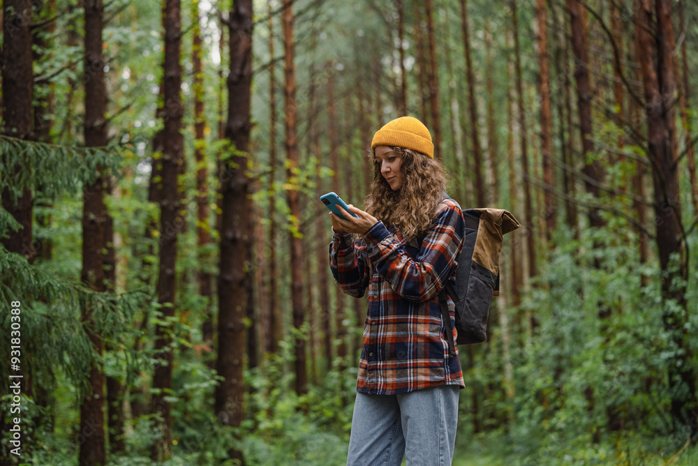 Fototapeta premium Young woman in a plaid shirt and beanie checking her smartphone while hiking through a forest, blending technology with outdoor adventure. 