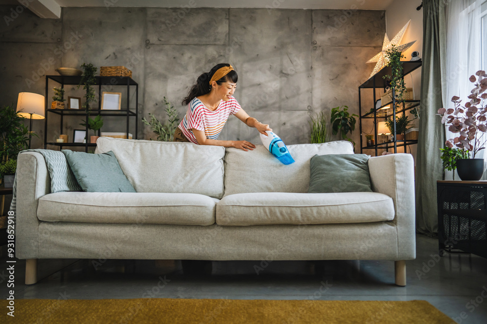 Obraz premium Mature japanese woman vacuums sofa with a hand vacuum cleaner at home