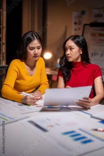 Two young asian businesswomen working late and discussing documents in office at night