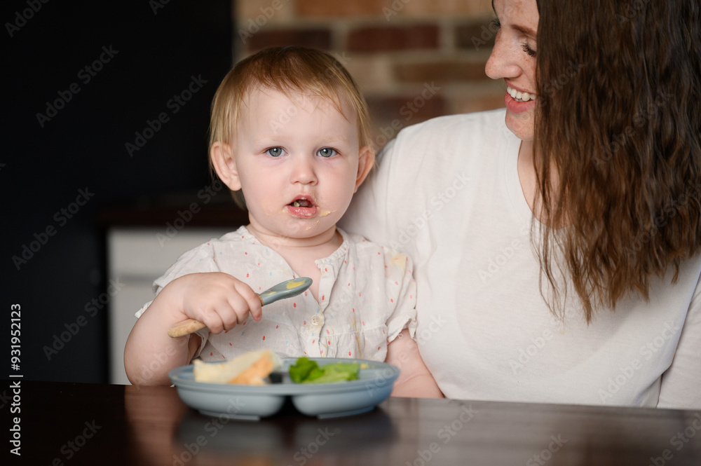 Happy smiling mother looking at baby eating puree from plate on her own while sitting on mom lap in kitchen at home. Toddler enjoying tasty puree. Baby feeding on her own, healthy food, weaning Happy smiling mother looking at baby eating puree from plate on her own while sitting on mom lap in kitchen at home. Toddler enjoying tasty puree. Baby feeding on her own, healthy food, weaning
