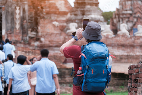 Tourists carrying backpacks and hats stand and take photos of an old temple with their smartphones.