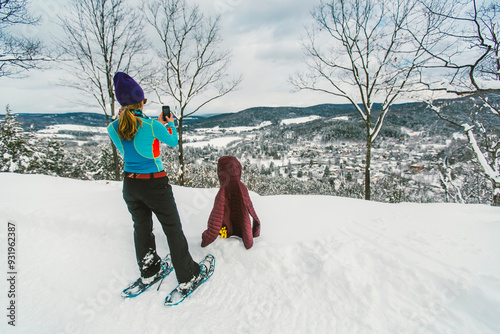 Woman snowshoeing in fresh snow stopping to take a cell phone picture of a small Vermont town from a top a mountain.