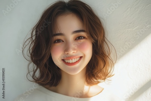 Close-up portrait of a smiling modern fashion model with shoulder-length dark hair, wearing a light-colored top, standing against a light background.