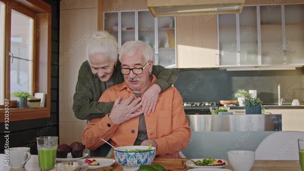 Medium shot of senior Caucasian man sitting at table, having healthy lunch, using smartphone, loving wife surprising husband with freshly baked muffins for desert, both hugging and smiling Medium shot of senior Caucasian man sitting at table, having healthy lunch, using smartphone, loving wife surprising husband with freshly baked muffins for desert, both hugging and smiling