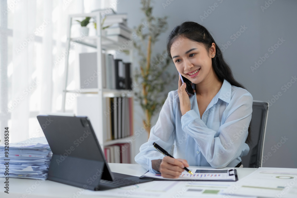 Young asian businesswoman working at her desk in a bright office, talking on the phone and taking notes on documents Young asian businesswoman working at her desk in a bright office, talking on the phone and taking notes on documents