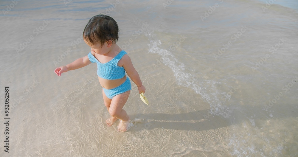 A young Toddler child wearing a blue swimsuit stands in shallow beach water, casting a shadow on a sunny day summertime