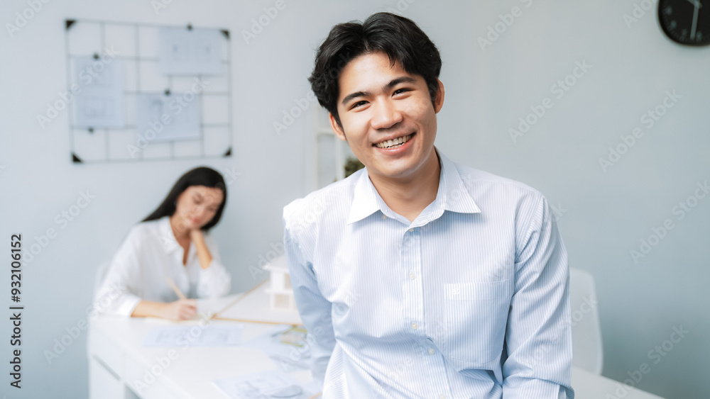 Professional happy businessman smiles during looking at camera. Young beautiful caucasian colleague drawing a blueprint on meeting table with house model and blueprint at modern office. Immaculate. Professional happy businessman smiles during looking at camera. Young beautiful caucasian colleague drawing a blueprint on meeting table with house model and blueprint at modern office. Immaculate.