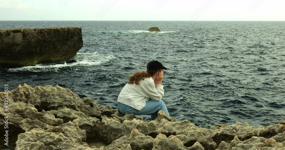 Depressive teen girl looks at azure waves leaning head on hands sitting on rocks at ocean resort. Teenager kid sits thinking on ocean beach on summer vacation Depressive teen girl looks at azure waves leaning head on hands sitting on rocks at ocean resort. Teenager kid sits thinking on ocean beach on summer vacation