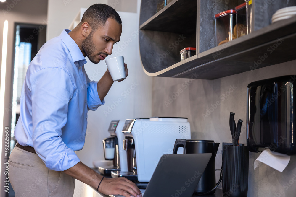 Obraz premium African American man wearing blue shirt working on laptop in kitchen
