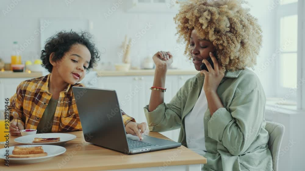 African American woman sitting at kitchen table, talking on cell phone and working on laptop during breakfast with little son eating jam toast beside her in the morning at home African American woman sitting at kitchen table, talking on cell phone and working on laptop during breakfast with little son eating jam toast beside her in the morning at home