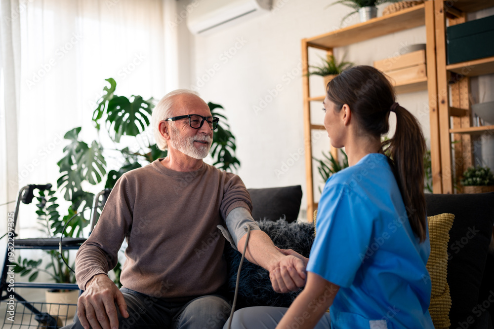 Obraz premium A cheerful nurse in blue scrubs takes the blood pressure of an elderly male patient during a home visit, capturing a moment of attentive health care service.