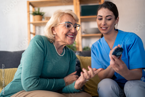 A smiling female nurse in blue scrubs attentively measures the blood glucose level of a happy elderly woman at her cozy apartment, ensuring meticulous in-home patient care. photo