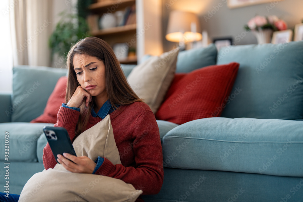A pensive Caucasian female clutches a pillow while browsing her smartphone, seated on a comfortable couch, her casual home attire adding to the intimate indoor setting. A pensive Caucasian female clutches a pillow while browsing her smartphone, seated on a comfortable couch, her casual home attire adding to the intimate indoor setting.