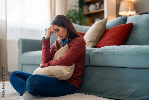 A Caucasian woman appears troubled, clutching a pillow while seated on the floor by a couch in a well-kept living room.