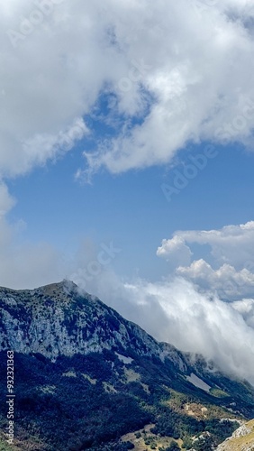 A dramatic scene of a mountain peak covered in dense greenery, partially hidden by swirling clouds under a bright blue sky.