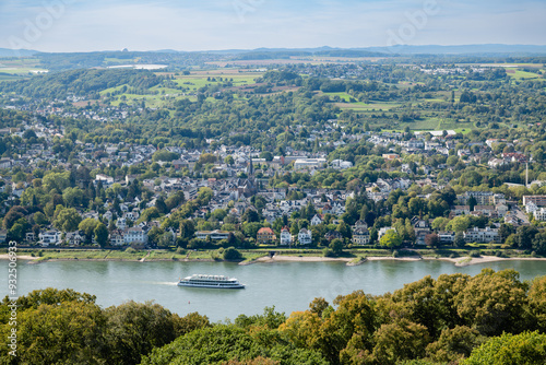 Scenic view of the Rhine River with a cruise ship passing by, framed by lush greenery and countryside buildings along the riverbank, under a clear blue sky.