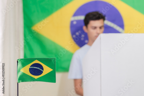 Bandeira brasileira decorando local de votação. No segundo plano, há um eleitor  desfocado escolhendo o seu candidato na urna eletrônica. Eleições municipais e presidenciais no Brasil  photo