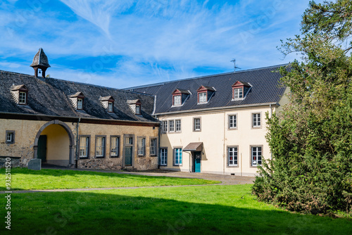 A yellow building with a slate roof and an arched entrance, located within the historical grounds of Klosterruine Heisterbach, framed by green lawns.