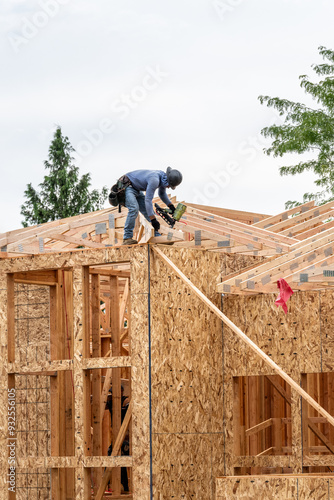 New residential home construction in framing stage, workman on the roof rafters working with pneumatic nail gun, OSB, oriented strand board, sheathing on exterior walls with window holes cut out 