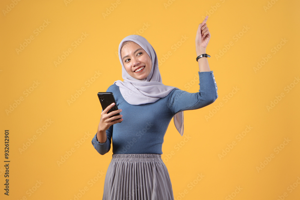 happy asian indonesian muslim woman holding smart phone with pointing finger gesture on isolated yellow background happy asian indonesian muslim woman holding smart phone with pointing finger gesture on isolated yellow background