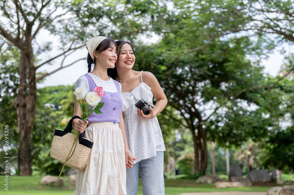Two beautiful Asian women are holding hands and looking away while standing together in a park. Two beautiful Asian women are holding hands and looking away while standing together in a park.