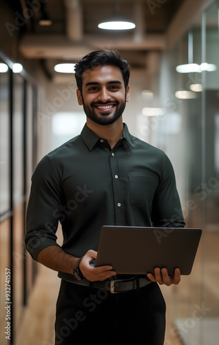 an attractive indian male social worker in his late thirties, smiling and holding a laptop while standing inside the office hallway
