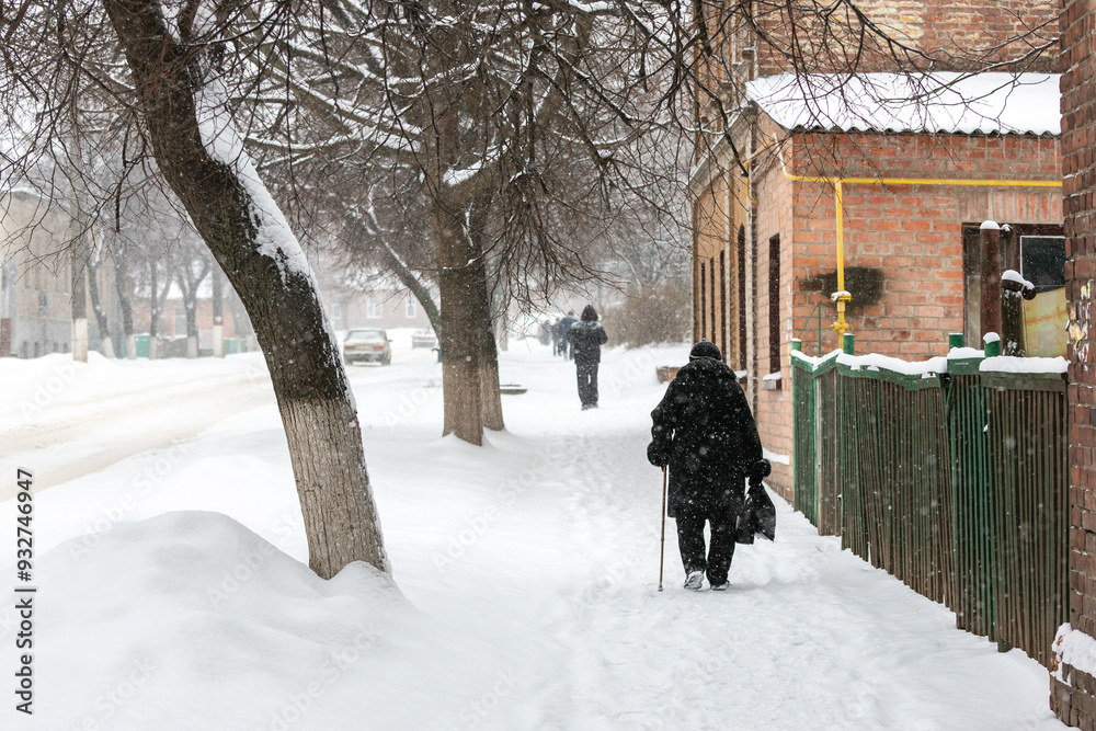 An old grandmother with a stick walks down a winter street during a snowfall. A woman walks in the snow on the street. Pensioner in the city in winter. Rear view An old grandmother with a stick walks down a winter street during a snowfall. A woman walks in the snow on the street. Pensioner in the city in winter. Rear view
