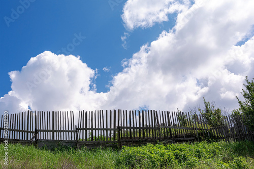 A rustic scene of an old wooden fence against a backdrop of a blue sky with fluffy clouds.