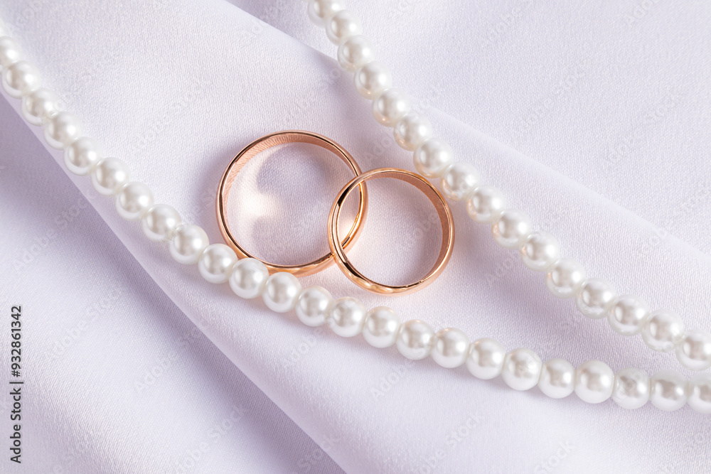 Two elegant gold wedding rings lie on a large fold of white satin among pearl strings of beads. top view. The concept of the wedding. postcard. Two elegant gold wedding rings lie on a large fold of white satin among pearl strings of beads. top view. The concept of the wedding. postcard.