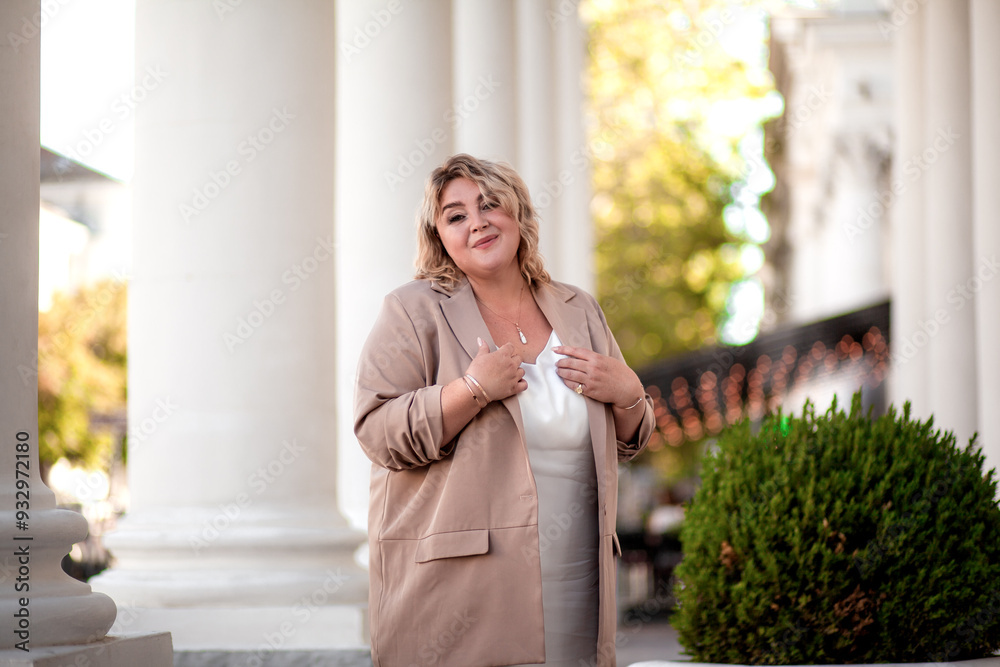 Fototapeta premium A beautiful fat middle aged woman walks around the city in a stylish image. The plus size model is dressed in a biege jacket and a white dress