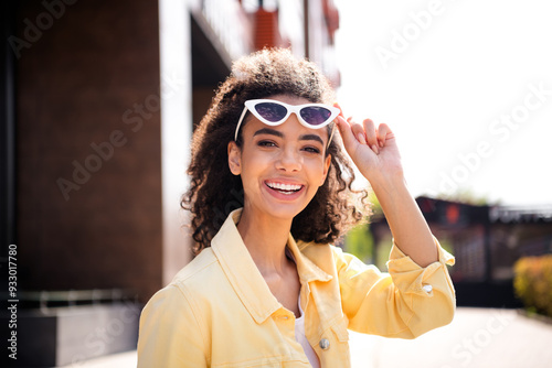 Photo of young joyful girl take off sunglasses with wavy hair student in yellow stylish jacket spending time outdoors sunny weather